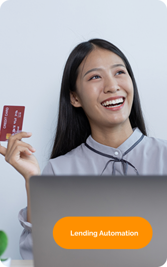 A person sits at a desk with a laptop, smiling and holding a smartphone in one hand and a credit card in the other. A small plant is placed nearby. The background is a plain white wall.